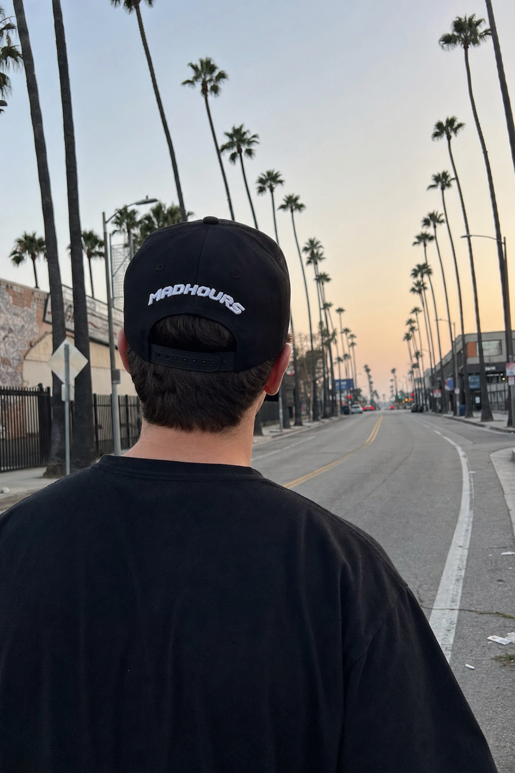 Rear view of the MADHOURS wordmark embroidered on a black snapback on a palm-lined Los Angeles street at dusk